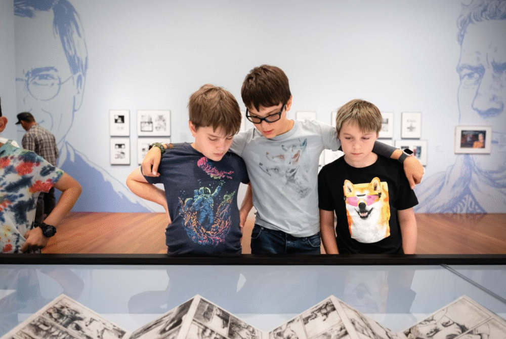 3 boys looks at manga in a glass case, middle boy with arms around the others