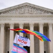 flags waving in front of the supreme court building one rainbow on top and one trans flag on bottom with a flag that says "trans Equality now"