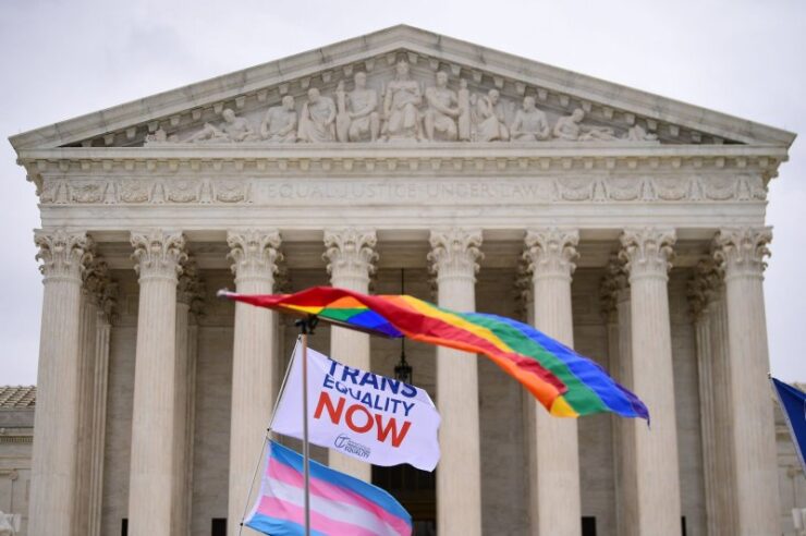 flags waving in front of the supreme court building one rainbow on top and one trans flag on bottom with a flag that says "trans Equality now"