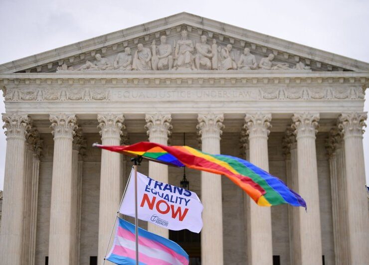 flags waving in front of the supreme court building one rainbow on top and one trans flag on bottom with a flag that says "trans Equality now"