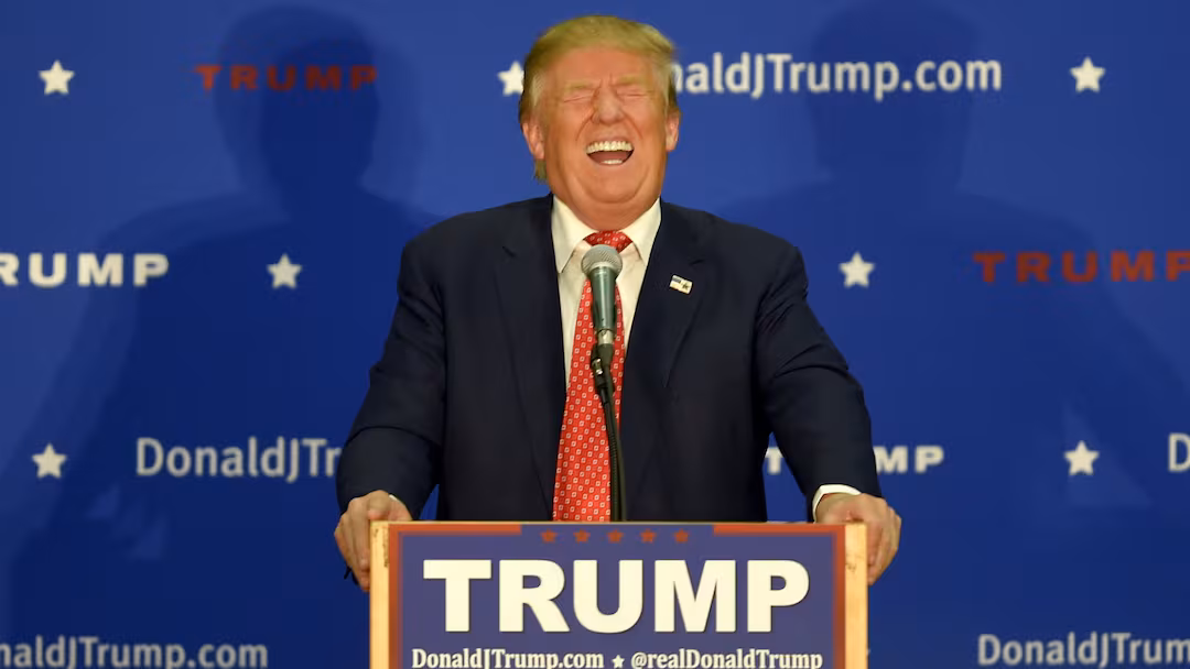 Trump laughing hysterically behind a podium with his name on it