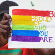 Citizen presumably of Uganda holding a sign that is rainnbow each color leading into the words "Be proud of who you are"